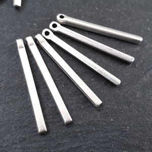 Puede incluir: Un grupo de seis colgantes de barra de metal plateado con un bucle en la parte superior. Los colgantes están dispuestos en forma de abanico sobre una superficie gris oscura.