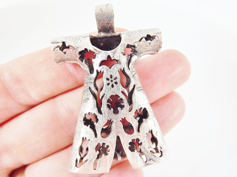 Pu&ograve; includere: Ciondolo color argento con un design floreale stilizzato. Il ciondolo ha una finitura strutturata e presenta forme floreali e di foglie ritagliate con accenti rossi e rosa. Il ciondolo &egrave; tenuto in mano su sfondo bianco.