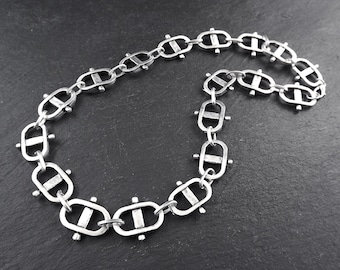 May include: A silver chain necklace with a unique link design. Each link is oval-shaped with a horizontal bar across the center and small round accents on the sides.