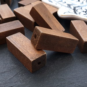 Puede incluir: Un grupo de cuentas de madera marrón con agujeros perforados en el centro. Las cuentas están dispuestas en una pila sobre una superficie gris oscuro.