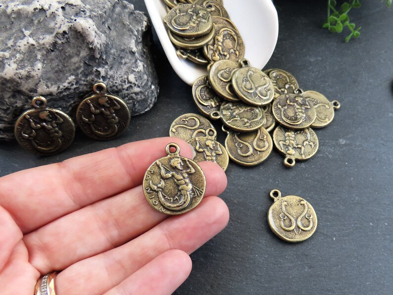 May include: A collection of antique gold-toned metal charms featuring a mermaid design. The charms are round and have a detailed design of a mermaid with flowing hair and a tail.