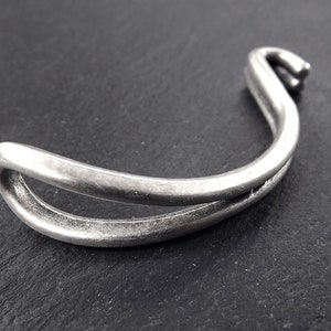 Può includere: Un braccialetto in metallo argentato con un design curvo e aperto. Il braccialetto ha una finitura liscia e lucida.