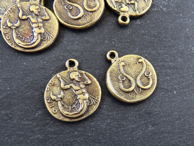 May include: Two gold-toned metal charms featuring zodiac signs. One charm depicts a mermaid with the word "CAPRICORN" inscribed around the edge. The other charm depicts two snakes intertwined with the word "SERPENS" inscribed around the edge.