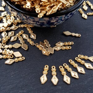 May include: A bowl filled with gold-colored metal charms with a diamond shape and beaded accents. The charms are scattered on a black surface.
