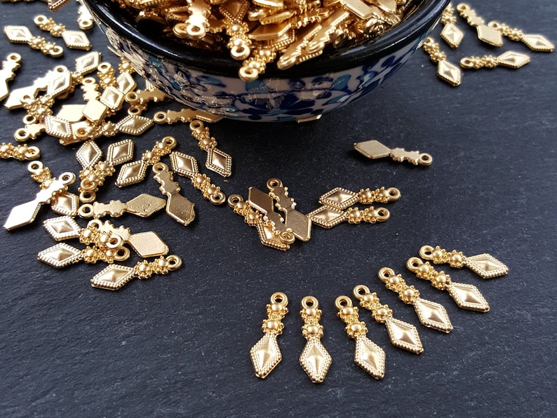 May include: A bowl filled with gold-colored metal charms with a diamond shape and beaded accents. The charms are scattered on a black surface.