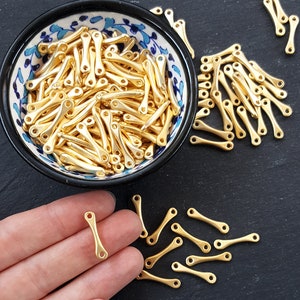 May include: A bowl filled with gold-colored metal connector pieces. The pieces are shaped like a long, thin rectangle with a small loop at each end. The bowl is decorated with blue and white floral patterns. A hand is holding one of the connector pieces.
