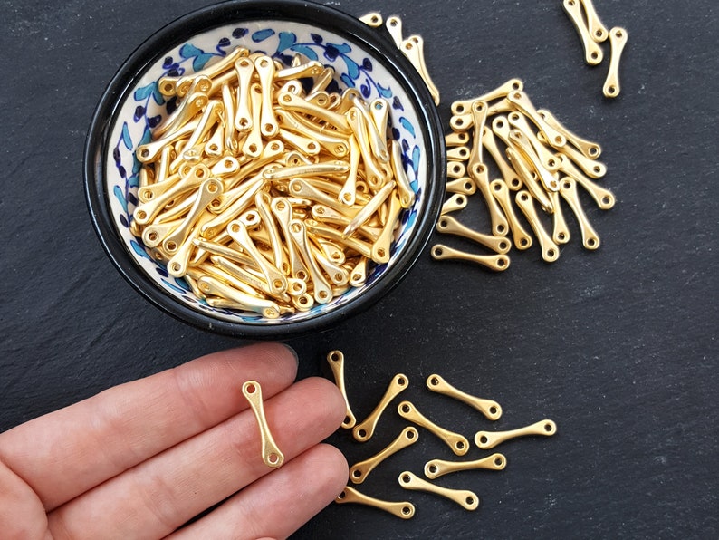 May include: A bowl filled with gold-colored metal connector pieces. The pieces are shaped like a long, thin rectangle with a small loop at each end. The bowl is decorated with blue and white floral patterns. A hand is holding one of the connector pieces.