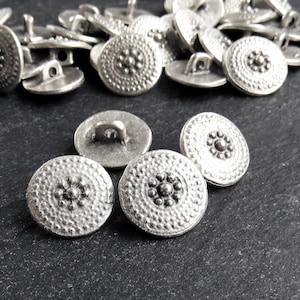 Könnte beinhalten: Eine Sammlung silberfarbener, runder Knöpfe mit strukturierter, gehammerter Optik. Jeder Knopf hat ein zentrales, erhabenes Design, das von einem Ring kleinerer, strukturierter Details umgeben ist. Die Knöpfe haben eine Schlaufe auf der Rückseite zum Annähen.