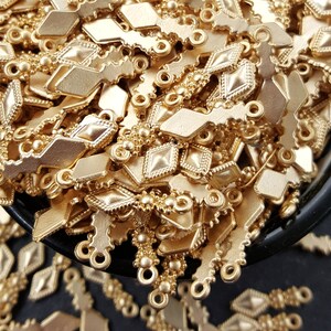 May include: A large pile of gold-colored metal charms in the shape of a diamond with a small loop at the top. The charms are arranged in a random pattern and are all the same size.