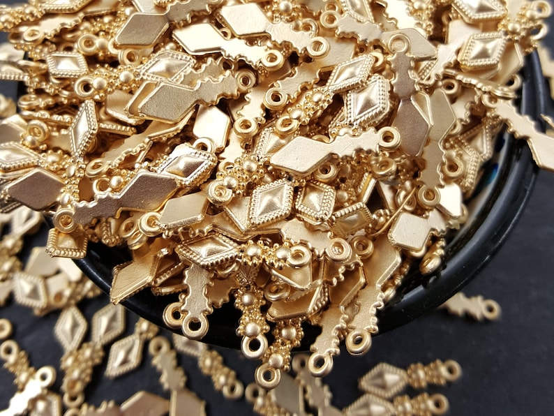 May include: A large pile of gold-colored metal charms in the shape of a diamond with a small loop at the top. The charms are arranged in a random pattern and are all the same size.