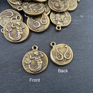 May include: A gold-toned metal charm with a detailed image of a mermaid on the front and a snake on the back. The charm has a loop for attaching to a necklace or bracelet.