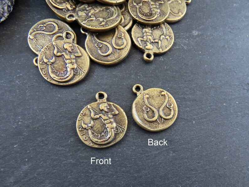 May include: A gold-toned metal charm with a detailed image of a mermaid on the front and a snake on the back. The charm has a loop for attaching to a necklace or bracelet.