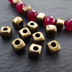 May include: A close-up of ten antique gold square beads with a hole in the center. The beads are scattered on a dark gray surface.