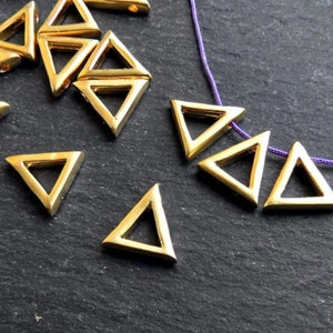 May include: A group of gold triangle-shaped beads with a hole in each corner. The beads are scattered on a dark gray surface. Some of the beads are strung on a purple cord.