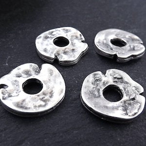 Puede incluir: Cuatro anillos de metal plateados con textura martillada y un agujero en el centro. Los anillos están dispuestos en semicírculo sobre una superficie gris oscura.