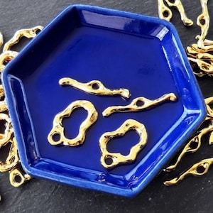 May include: A close-up of a blue ceramic dish filled with gold-colored metal charms. The charms are in various shapes, including circles, ovals, and abstract shapes. Some of the charms have a smooth finish, while others have a textured finish.
