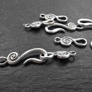 May include: A collection of silver-coloured metal clasps featuring a decorative swirl design. The clasps vary in size and shape, with a hook and eye closure. The background is a dark, textured surface.