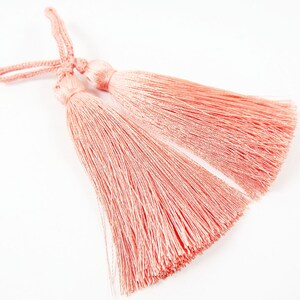 May include: Two pink tassel trims with a knotted cord. The tassels are made of soft, fine fibers and are approximately 2 inches long.