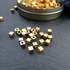 May include: A collection of small, gold-colored, square beads with a hole through the center. Some beads are scattered on a dark surface, while others are in a small, round, metal container. The beads appear to be made of metal.