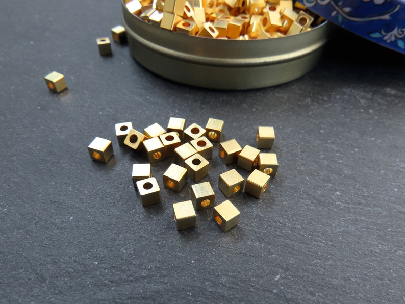 May include: A collection of small, gold-colored, square beads with a hole through the center. Some beads are scattered on a dark surface, while others are in a small, round, metal container. The beads appear to be made of metal.