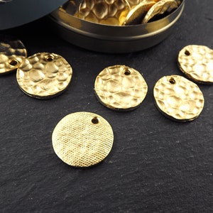 Może przedstawiać: Zbliżenie kilku złotych, teksturowanych metalowych zawieszek. Zawieszki są okrągłe, z młotkowaną lub tkaną fakturą i małym otworem u g&oacute;ry. Niekt&oacute;re zawieszki są rozrzucone na ciemnej powierzchni, inne w małym, otwartym pudełku.