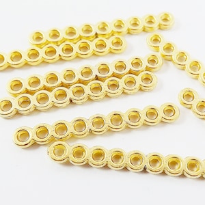 Op de afbeelding: Een set van acht gouden metalen connectorstukken, elk met een rij van acht kleine, open cirkels. De stukken zijn verspreid op een wit oppervlak.