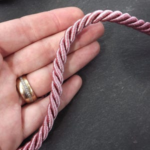 May include: A close-up of a pink braided cord, showing the texture and detail of the cord. The cord is held in a hand, with a ring on the finger.