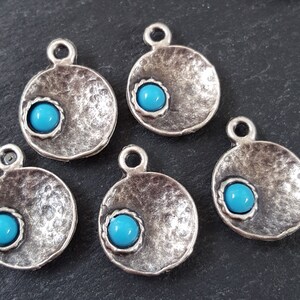 May include: Five silver-toned round pendants with a turquoise blue cabochon stone in the center of each. The pendants have a textured surface and a loop at the top for hanging.