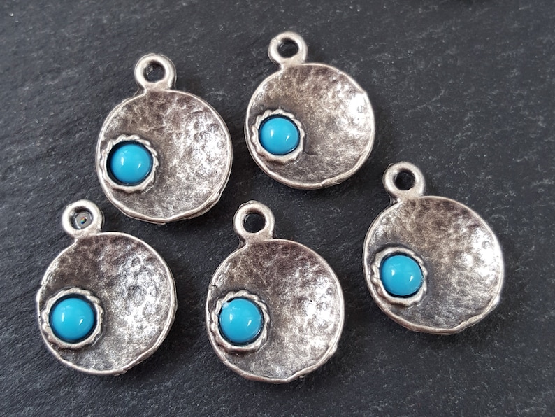 May include: Five silver-toned round pendants with a turquoise blue cabochon stone in the center of each. The pendants have a textured surface and a loop at the top for hanging.