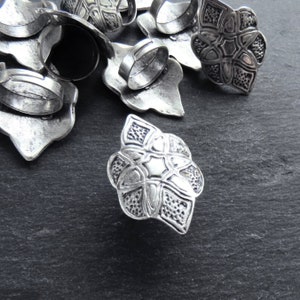 May include: Silver-toned metal rings with ornate floral designs. The rings feature adjustable bands and detailed, textured flower-shaped faces. The rings are scattered on a dark, textured surface, showcasing their intricate craftsmanship.