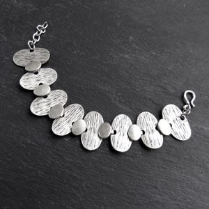 May include: A silver bracelet with a chain and a clasp. The bracelet is made of a series of oval-shaped links, each with a textured surface.