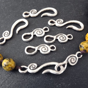 May include: A selection of silver-coloured jewellery clasps with a swirling pattern are shown. The clasps have different sizes and shapes, some with a hook and others with a double-loop design. A beaded necklace with yellow and brown beads is also visible.