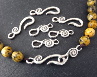 May include: Silver-toned jewelry clasps with a swirling design are displayed on a dark surface. The clasps vary in size and shape, with some featuring a hook and others a double-loop design. A beaded necklace with yellow and brown beads is also visible.