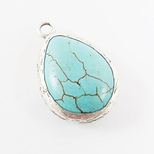 Op de afbeelding: Een druppelvormige hanger met een turquoise steen en een zilverkleurig metalen frame. De steen heeft een gebarsten patroon. De hanger heeft een kleine lus aan de bovenkant voor een ketting of koord.