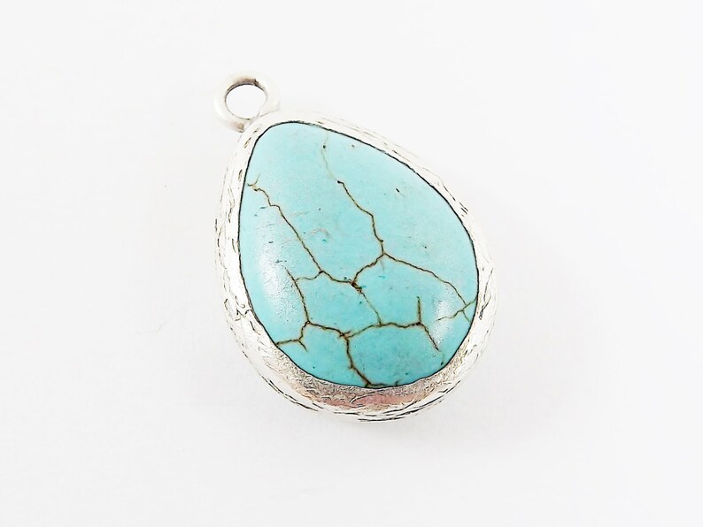 Op de afbeelding: Een druppelvormige hanger met een turquoise steen en een zilverkleurig metalen frame. De steen heeft een gebarsten patroon. De hanger heeft een kleine lus aan de bovenkant voor een ketting of koord.