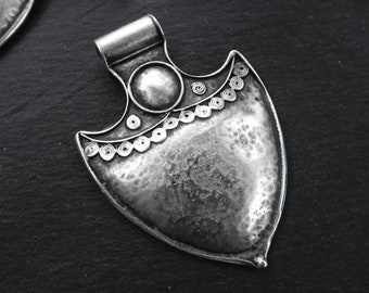 May include: A silver-toned pendant shaped like a shield with a textured surface and a circular design at the top. The pendant has a loop at the top for attaching a chain.