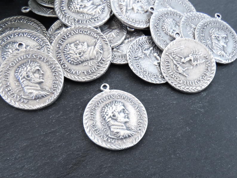 May include: A collection of silver-toned metal charms featuring Roman emperor Antoninus Pius. Each charm has a raised profile of the emperor's head and the inscription "ANTONINVS PIVS AVGGERM" around the edge.