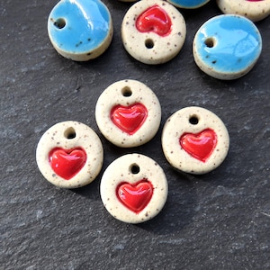 May include: A collection of ceramic beads. The beads are round with a speckled, off-white base. Each bead features a glossy red heart in the center. Some beads are also accented with a bright turquoise blue.