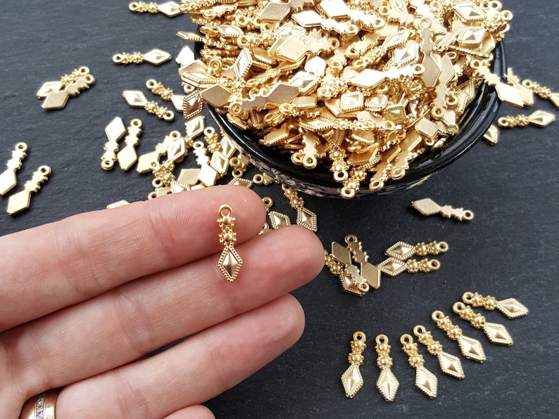 May include: Gold-toned metal pendant charms with a diamond shape and intricate details. The charms have a loop at the top for attaching to jewelry or other crafts.
