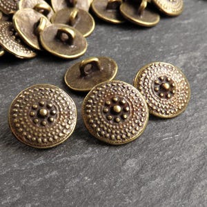May include: A collection of antique brass-coloured buttons. The buttons have a circular shape with a detailed, textured design. Each button features a central floral pattern and a loop on the back for sewing. They are scattered on a dark surface.