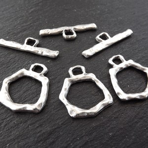 May include: Silver toggle clasps with a hammered finish. The clasps are in the shape of a rectangle with rounded corners. There are six clasps in total.