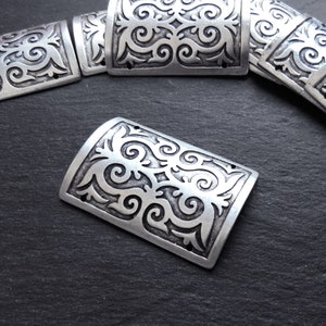 May include: Silver-toned metal embellishments with a decorative, swirling pattern. The embellishments are rectangular with rounded edges and have two small holes on each side.