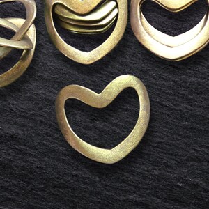 May include: Close-up of several brass-colored metal heart-shaped objects. One heart is a solid shape, while others are open. There is also a brass-colored metal knot-shaped object. The objects are on a dark, textured surface.