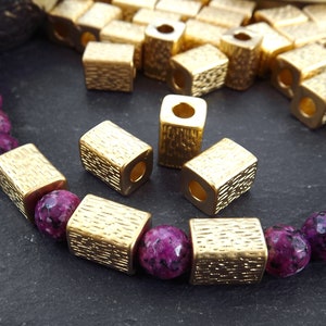May include: Gold metal spacer beads with a textured surface. The beads are square shaped with a hole in the center. The beads are shown on a strand of purple gemstone beads.