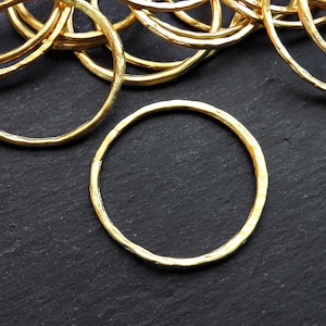 May include: A close-up of a pile of gold-colored metal rings. The rings are all the same size and have a hammered texture. The rings are on a black surface.