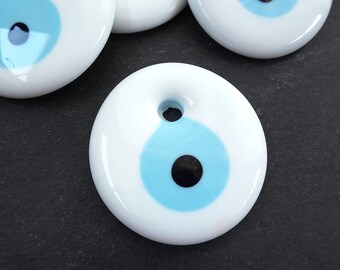 May include: White ceramic charm with a blue and black evil eye design. The charm has a hole for hanging.