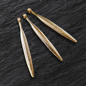 Könnte beinhalten: Drei goldfarbene Metall-Feder-Anhänger. Die Federn sind lang und schmal mit einer strukturierten Oberfläche.