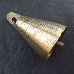 Op de afbeelding: Een kleine messing bel met een conische vorm en een lus aan de bovenkant om op te hangen. De bel heeft een getextureerd oppervlak met horizontale lijnen en een kleine, bruine klepel aan de binnenkant. De bel is ongeveer 3,8 cm lang.