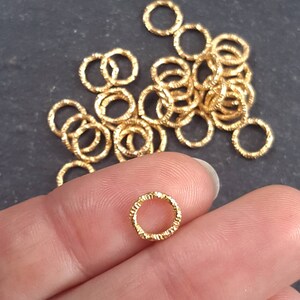 May include: A pile of gold-colored jump rings, some are textured, some are smooth. The rings are small and are used for jewelry making.