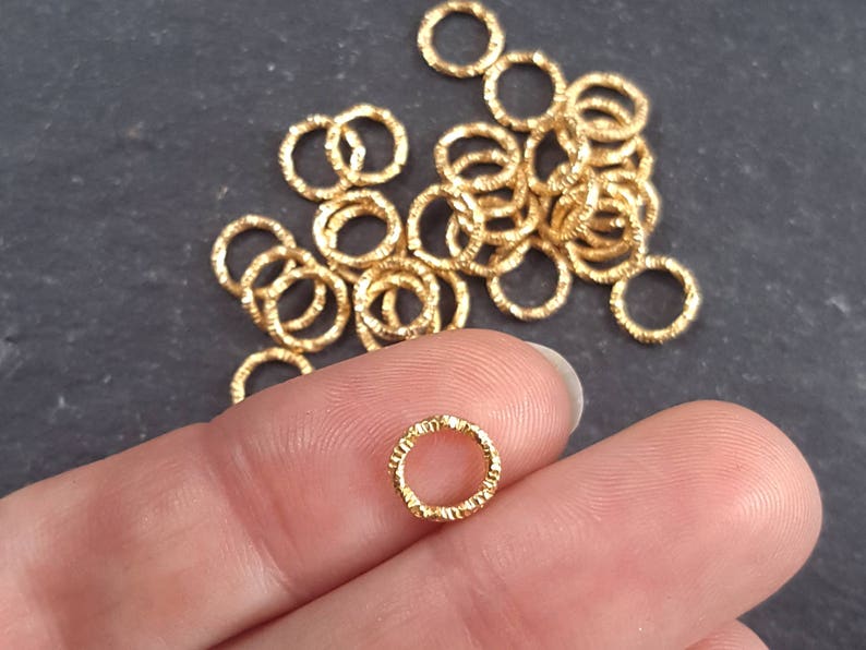 May include: A pile of gold-colored jump rings, some are textured, some are smooth. The rings are small and are used for jewelry making.
